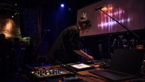 Contemporary live music stage with performers' instruments, controllers, microphones, monitors, lights, and blank screens